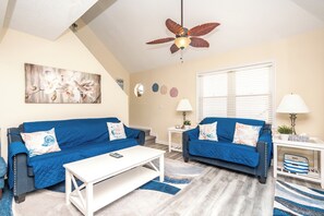 Ein Wohnzimmer mit zwei blauen Sofas, einem weißen Couchtisch, einem Deckenventilator und einem Fenster mit Jalousien.