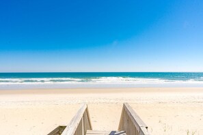 Une promenade en bois mène à une plage où l’eau est d’un bleu limpide et le ciel est dégagé.