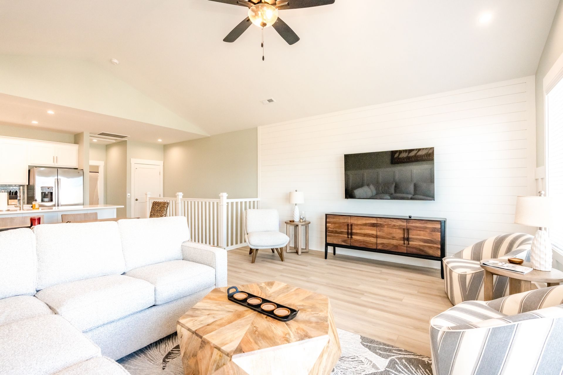 A modern living room with a sofa, a coffee table, a striped armchair, a dining area with a table and chairs, a kitchen with appliances, and a ceiling fan.
