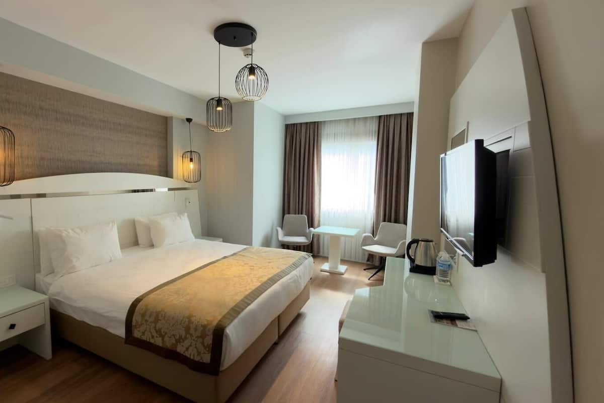 Quarto de hotel com cama, mesas de cabeceira, escrivaninha, cadeiras, televisão e janela com cortinas.