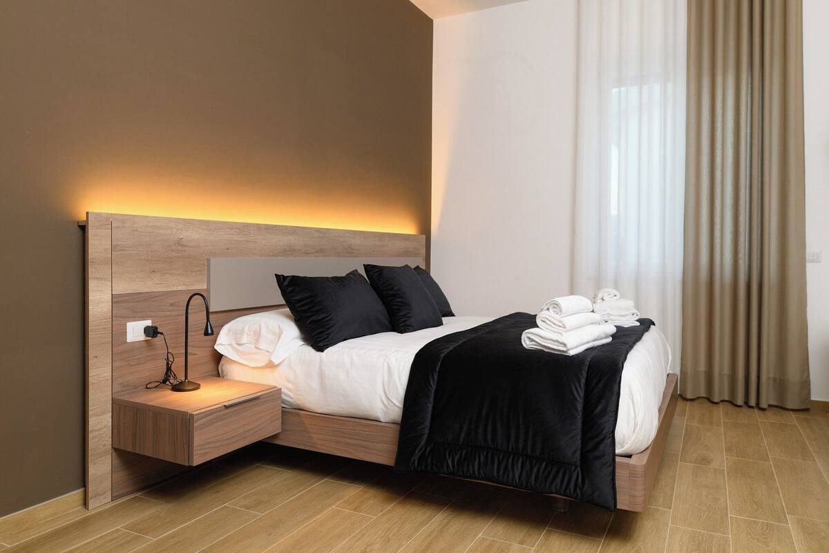 Een moderne hotelkamer met een houten hoofdeinde, een nachtkastje met een lamp, een opgemaakt bed met witte lakens en een zwarte plaid, en een raam met dunne gordijnen.