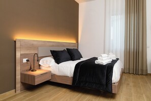 Et moderne hotelværelse med sengegavl i træ, et natbord med lampe, en pænt redt seng med hvide hør og et sort plaide, samt et vindue med lette gardiner.