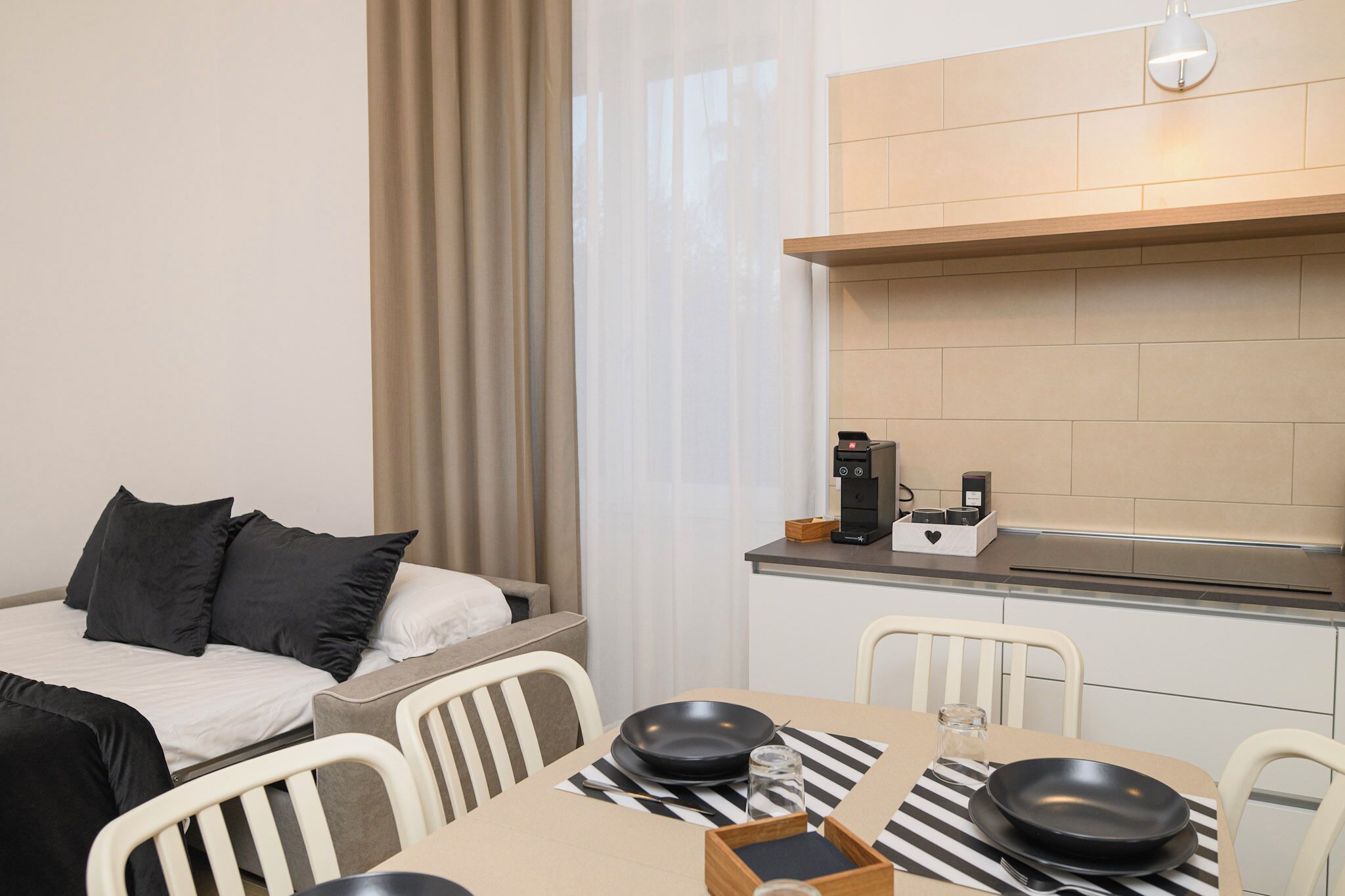 Ein kompaktes Hotelzimmer mit Bett, einer kleinen Küchenzeile, einem Essbereich mit Tisch und Stühlen sowie einem Fenster mit Vorhängen.