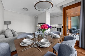 A modern dining area with a wooden table set for four, surrounded by grey upholstered chairs. A chandelier hangs above, and a kitchen is visible in the background. - Ochota Apt - Parking, Balcony & Railway Nearby (Warszawa)
