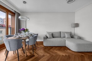 A modern living room with a grey sectional sofa, a dining table set for four, and a large window offering a view of buildings. - Ochota Apt - Parking, Balcony & Railway Nearby (Warszawa)