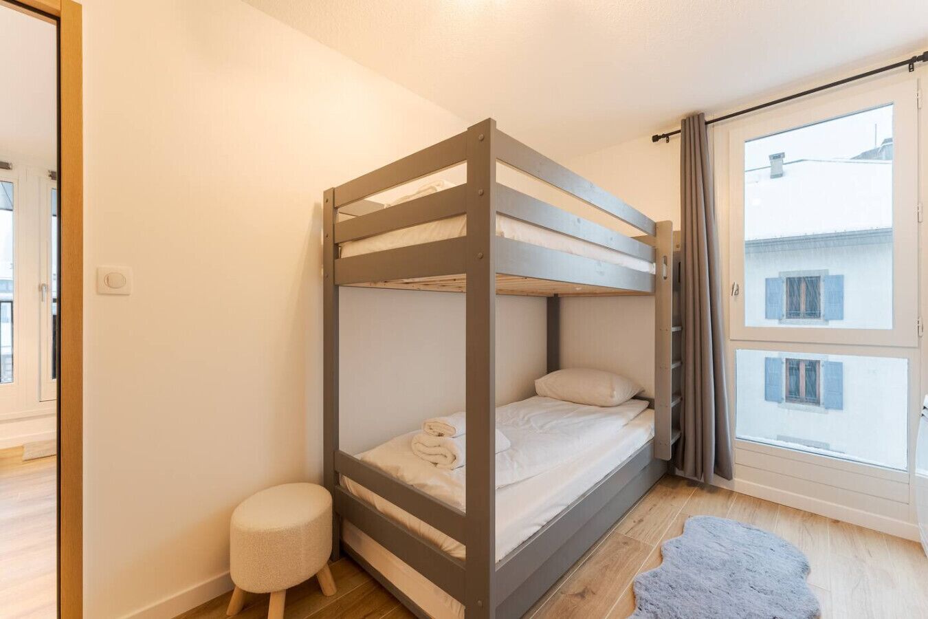 Ein Schlafraum mit Etagenbetten, einem kleinen Hocker, einem Fenster mit Vorhängen und Blick auf Gebäude.