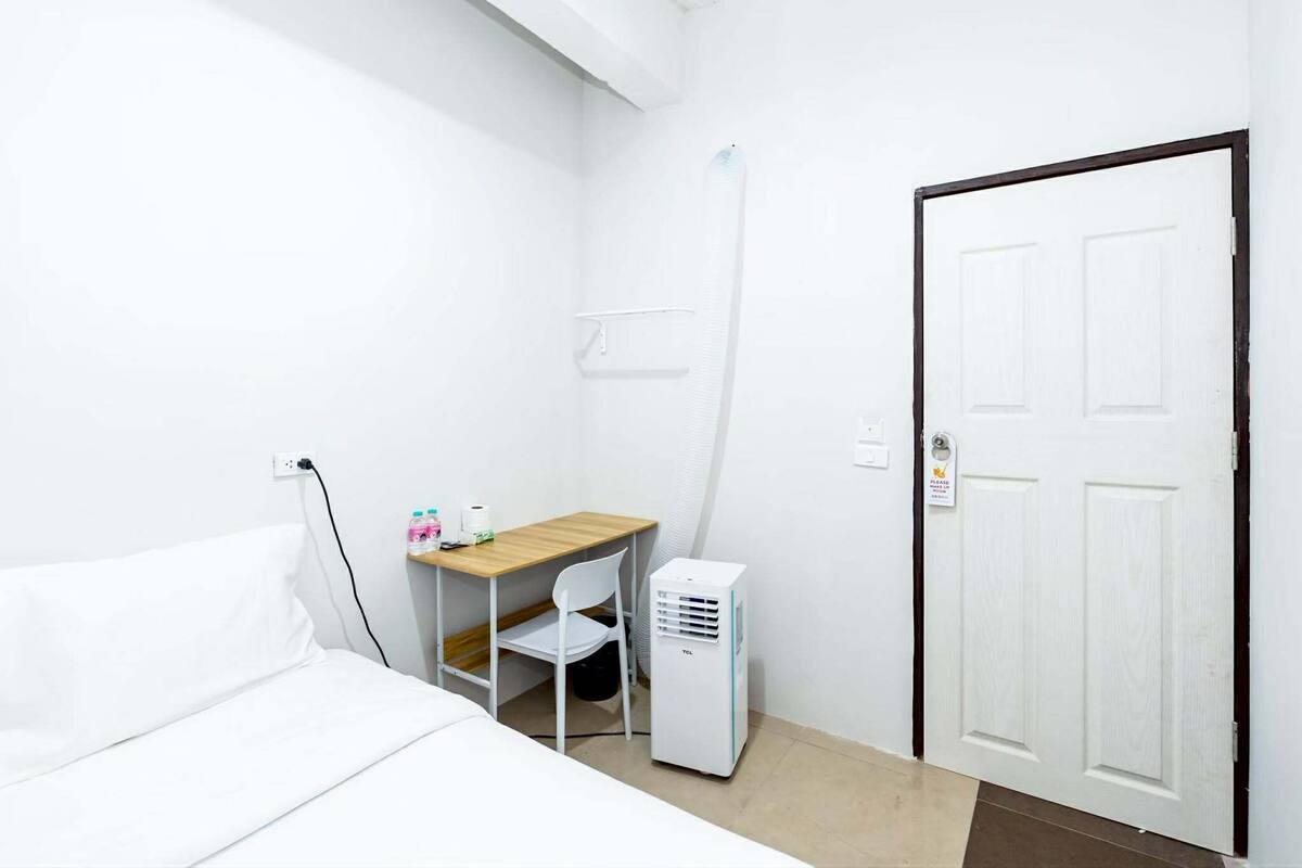A hotel room with a bed, a desk with a chair, a white air conditioner, and a door.
