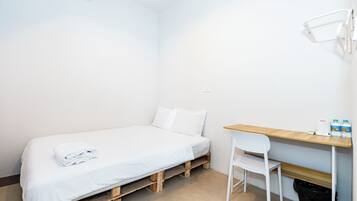Una habitación pequeña con una cama individual, paredes blancas, un escritorio de madera y una silla, un cubo de basura pequeño y una lámpara fijada a la pared.