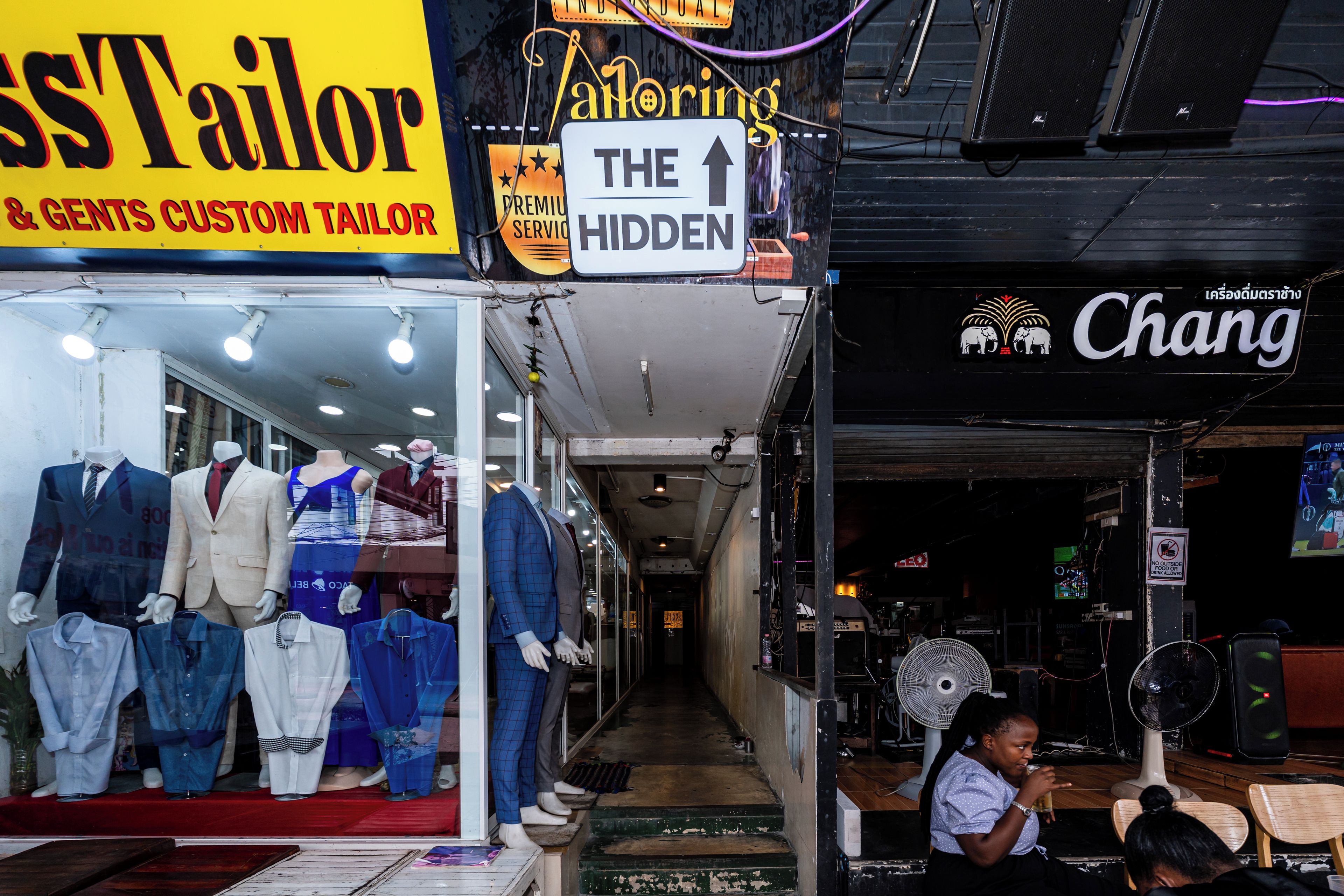 A narrow alley with shops, including a tailor shop with mannequins displaying suits and a sign that says 'THE HIDDEN'.
