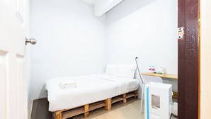 A small, white-walled room with a single bed, a wooden bench, a desk with a chair, and a white radiator. - The Hidden Khaosan (Bangkok)