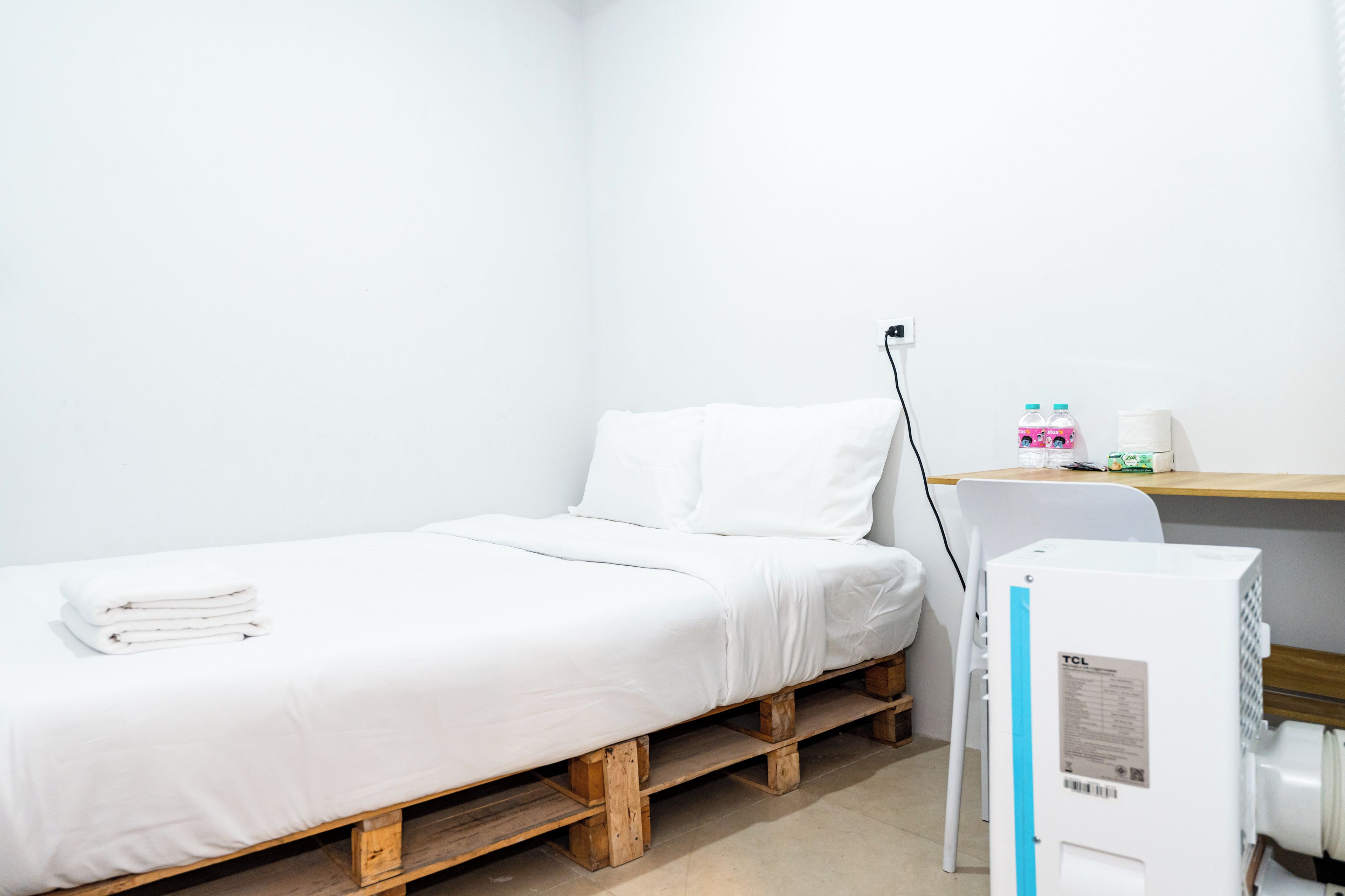 A small, single-bed room with a wooden bed frame, white bedding, and a desk with a computer and personal items.
