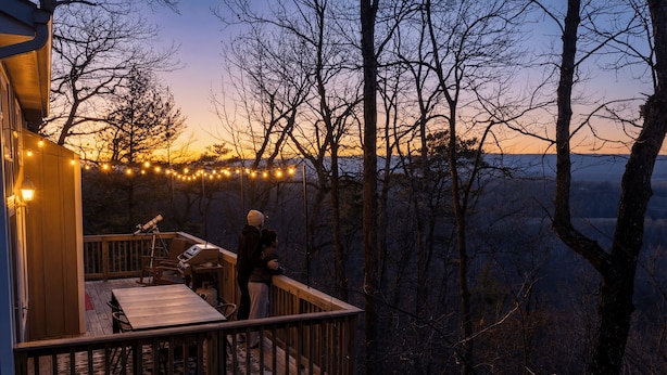 A wooden deck with a table and chairs, overlooking a forested area with string lights. - Country Road Cottage-Mountain Views + Private Hot Tub + Sauna + Game Room (Berkeley Springs)