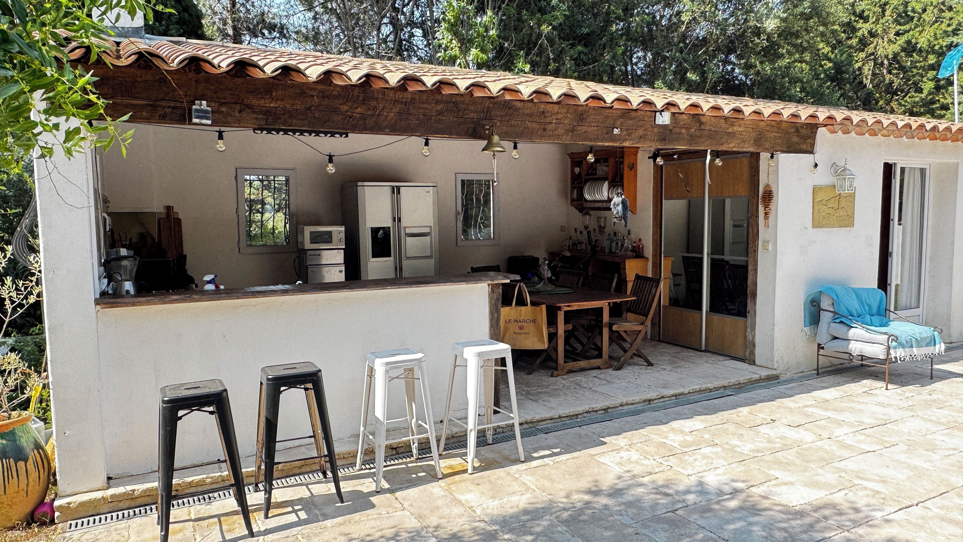 Une petite cuisine rustique avec un toit en tuiles, un coin repas avec une table et des chaises, et une chaise bleue à l’extérieur.