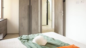 Een slaapkamer met een bed, een groene deken en een oranje kussen. Er is een kast en een deur naar een andere kamer.