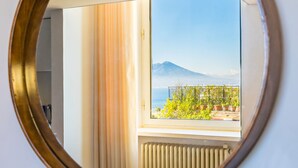 A round mirror with a gold frame reflecting a room with a window, a radiator, and a view of a mountain and sea. - Lite Life Panoramic Flat by Napoliapartments (Naples)