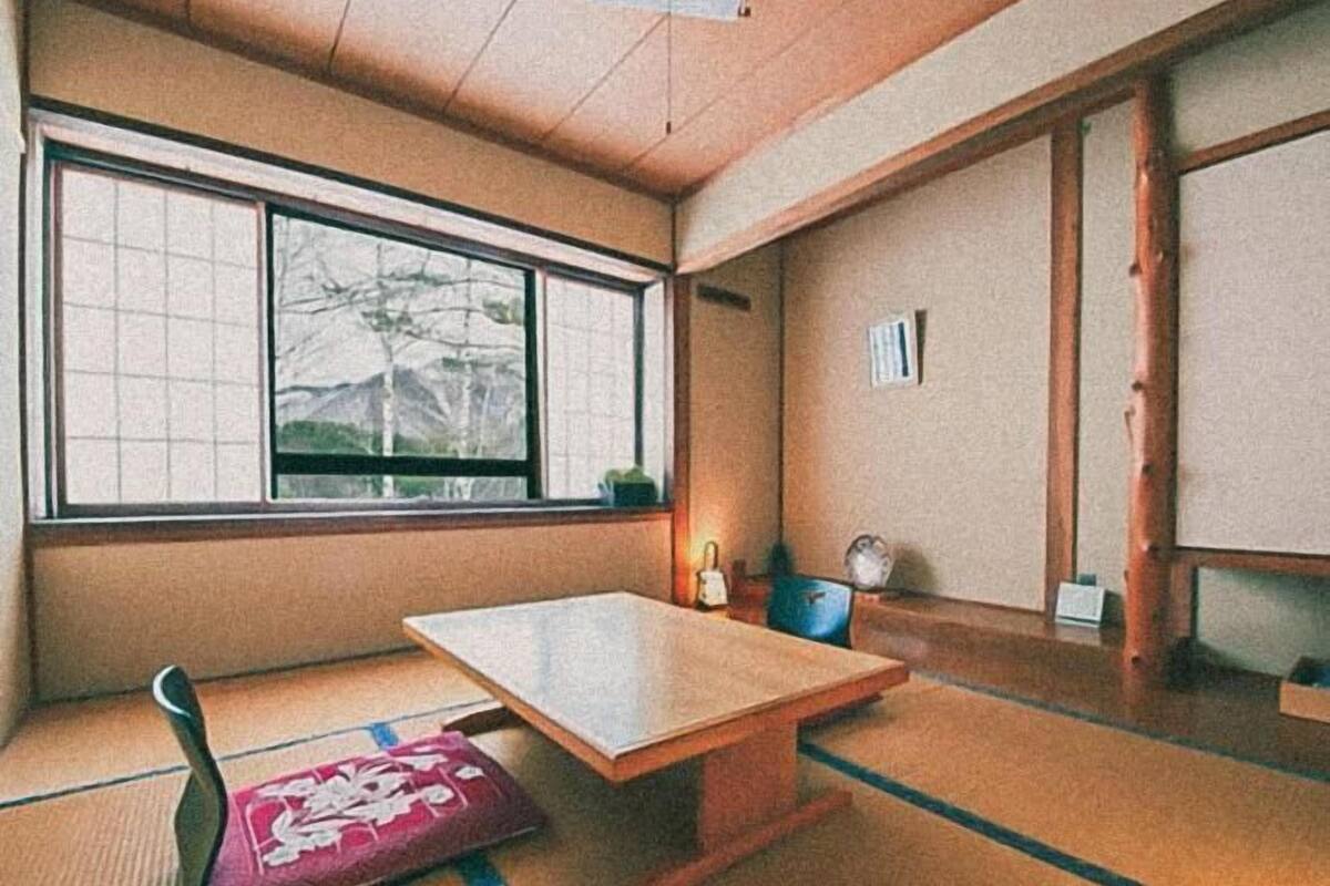A traditional Japanese room with tatami flooring, a low wooden table, a cushioned bench, and a large window offering a view of trees and mountains.