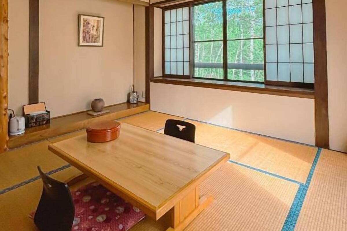 A traditional Japanese room with tatami flooring, a wooden table, and a large window with a view of greenery.