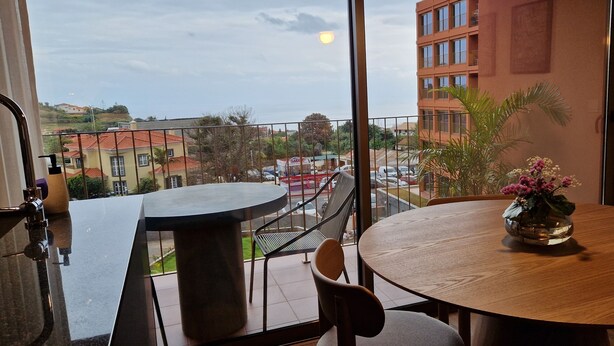 A balcony with a dining table and chairs, a flower vase, and a view of buildings and a street. - Shell Living - KOS (Funchal)