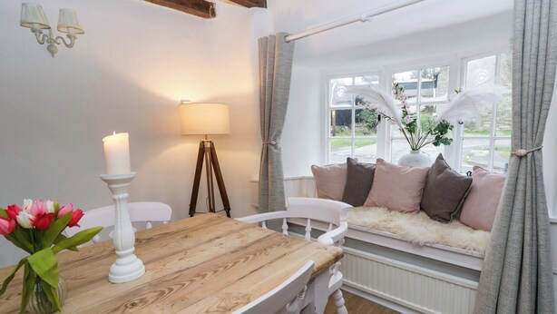 A cozy room with a wooden dining table, white chairs, a white bench with cushions, a standing lamp, and a window with curtains. - The Old Shop (Builth Wells)