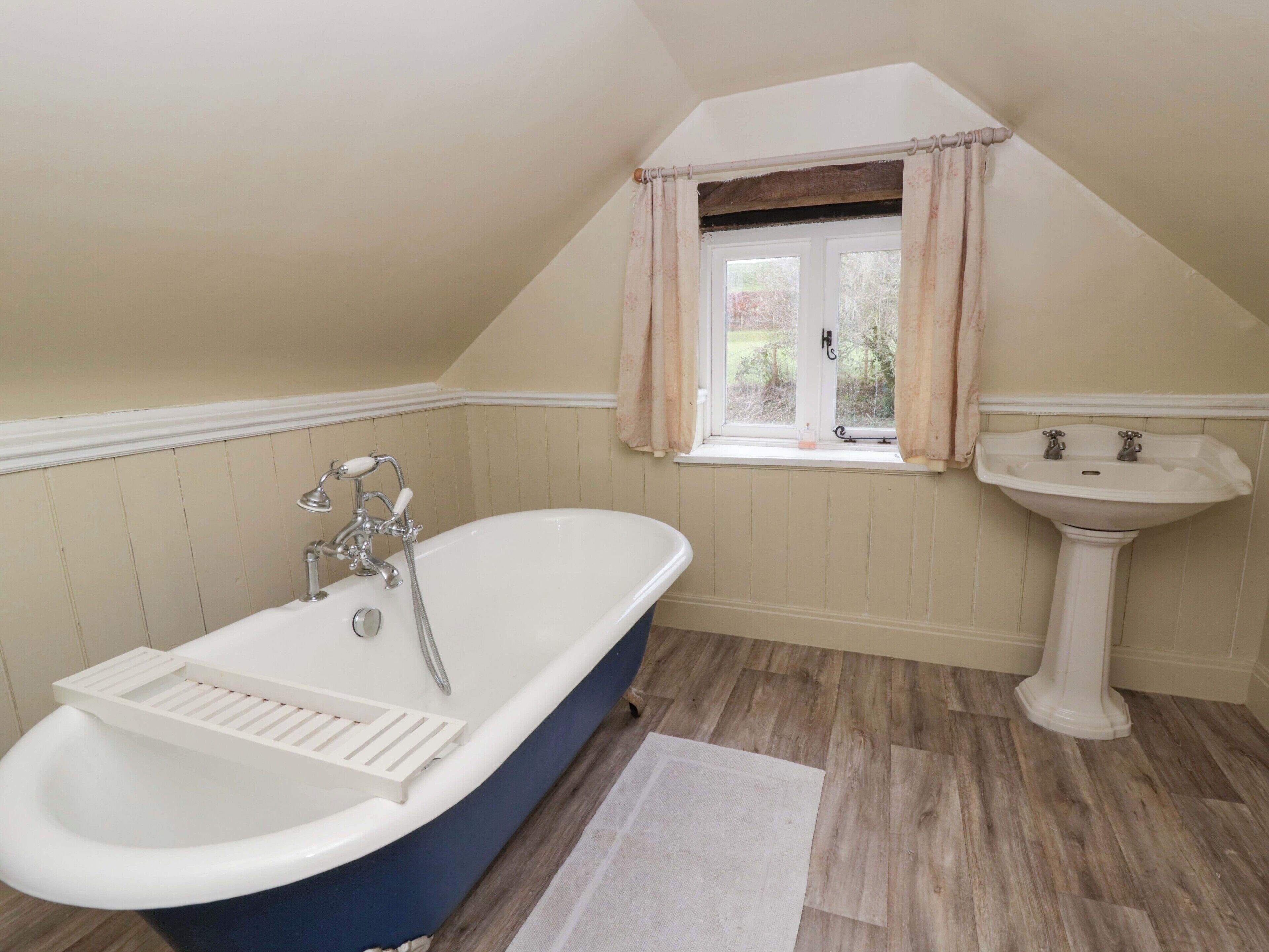 A bathroom with a freestanding bathtub, a window with curtains, a sink, and wooden flooring.