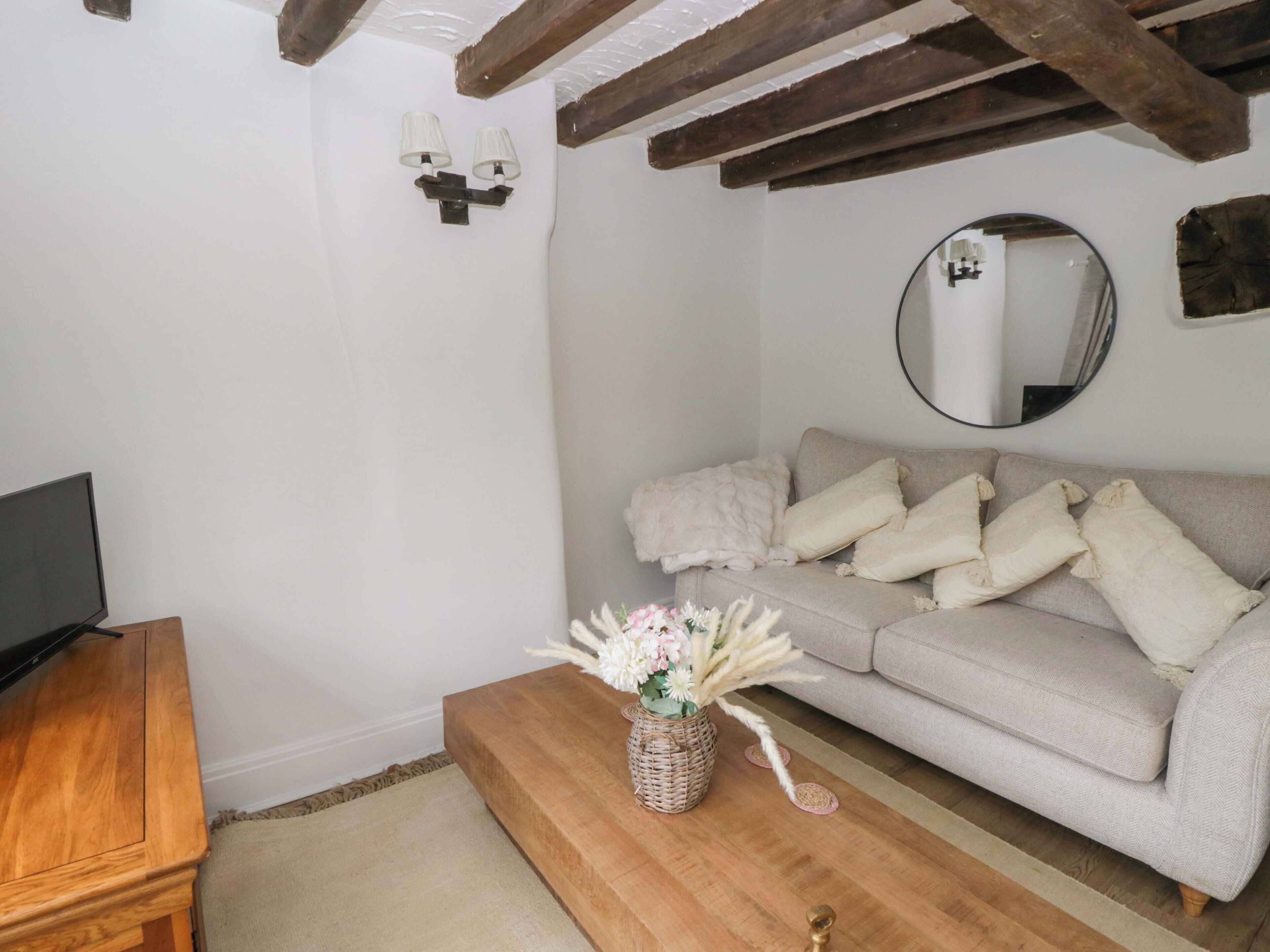 A living room with a beige sofa, a wooden coffee table with a vase of flowers, a flat-screen TV on a wooden stand, and a round mirror on the wall.