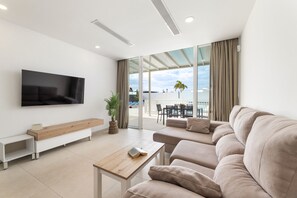 Una sala moderna con un televisor de pantalla plana grande, un banco de madera, un sofá color beige y un comedor con vistas al exterior.