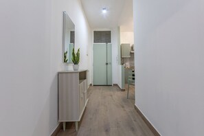 Un corridoio con pavimento in legno chiaro, parete bianca, uno specchio, una pianta in vaso e una porta verde chiaro, oltre a un angolo cottura.