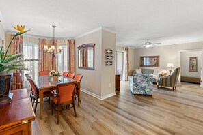 A spacious living room with a dining area, hardwood floors, a ceiling fan, and a large window with curtains. - Royale Palms 806 2 Br Oceanview w/Pool (Myrtle Beach)