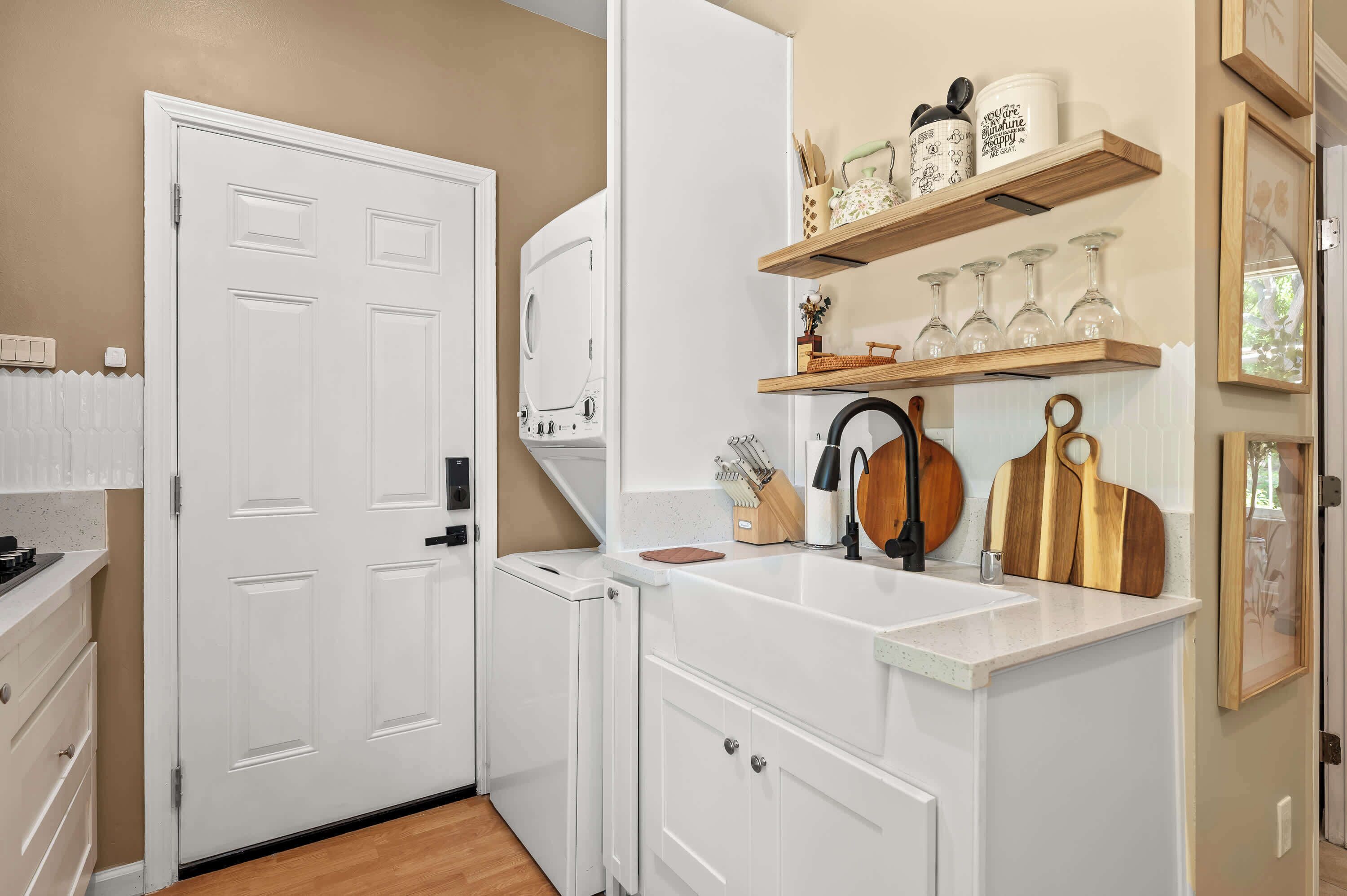 A kitchen with white cabinetry, a built-in oven, and a countertop with a sink. There are wooden shelves above the sink holding glassware and kitchen utensils.