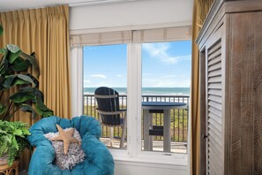 Una habitación con un amplio ventanal que da a una playa, un sillón azul decorado con una estrella de mar, y vistas al océano.