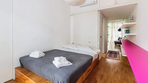 Een slaapkamer met een bed, een houten bankje, een witte airconditioning en een gang naar een woonruimte.
