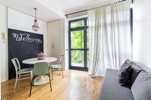 A modern living room with a sofa, a dining table with chairs, a blackboard with 'Welcome' written on it, and a large window with curtains. - Casa Watt Stylish Apartment With Balcony (Milan)
