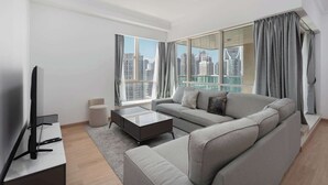 Ein modernes Wohnzimmer mit einem grauen Sofa, einem Couchtisch und einem Fernseher auf einem Schrank. Große Fenster bieten Blick auf eine Stadtlandschaft.