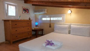 Une chambre avec une commode en bois, un lit recouvert de draps blancs, un climatiseur fixé au mur et une fenêtre agrémentée d’un rideau décoratif.