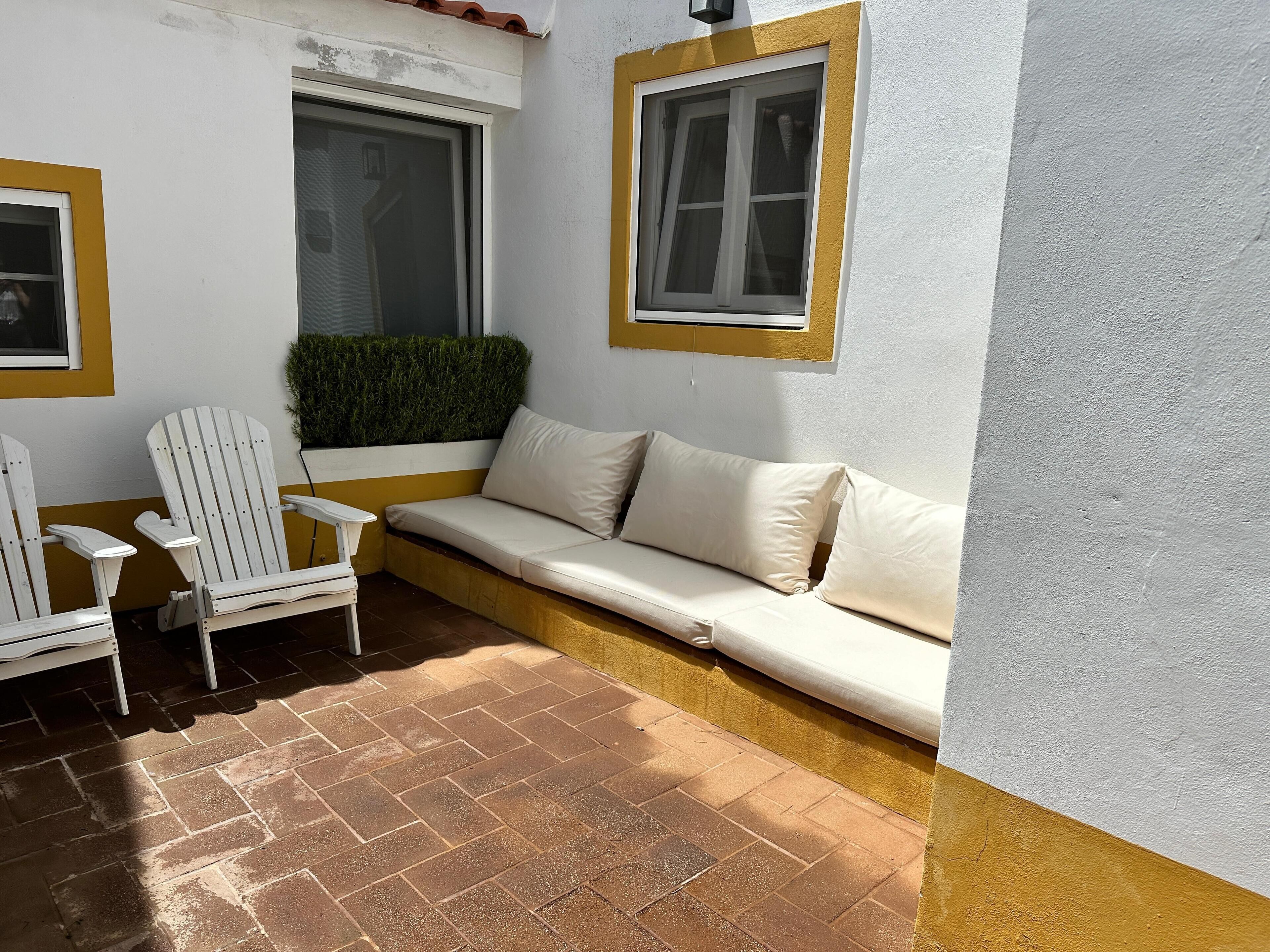 A corner seating area with a white sofa and two white Adirondack chairs, a small window with a yellow frame, and a brick floor.