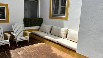 A corner seating area with a white sofa and two white Adirondack chairs, a small window with a yellow frame, and a brick floor.