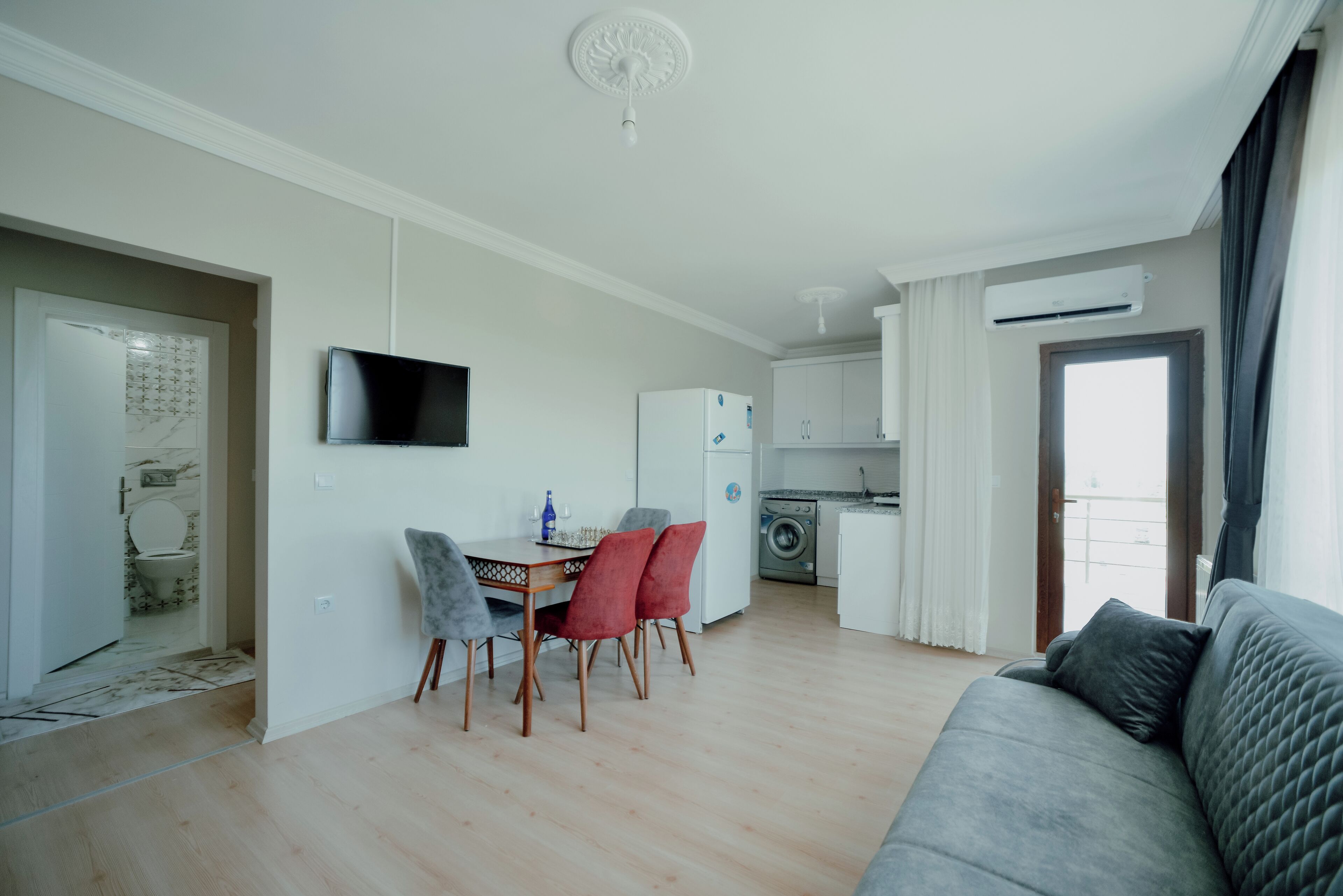 A modern living room with a grey sofa, a dining area with a table and chairs, a kitchen with a washing machine, and a TV mounted on the wall.