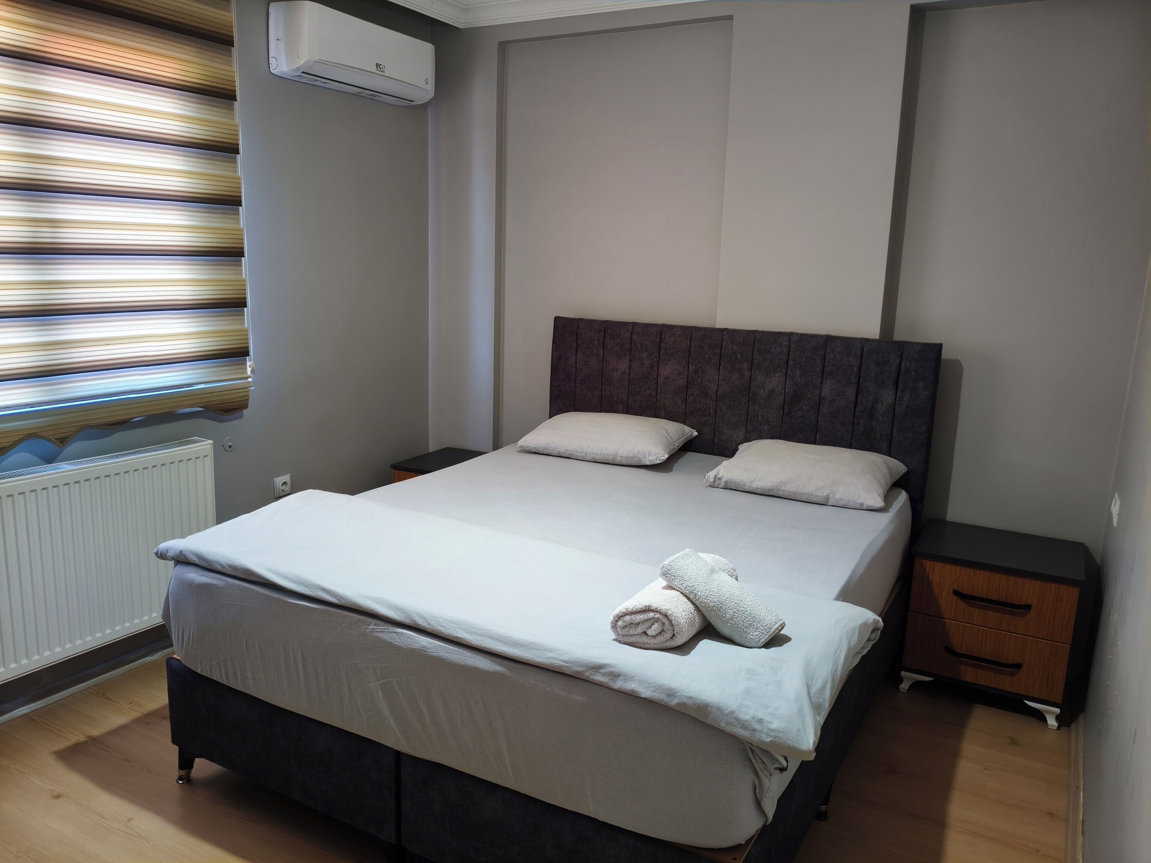 Een netjes opgemaakt bed met een witte deken en kussens, een houten nachtkastje met een lade, een radiator en een raam met zonwering.