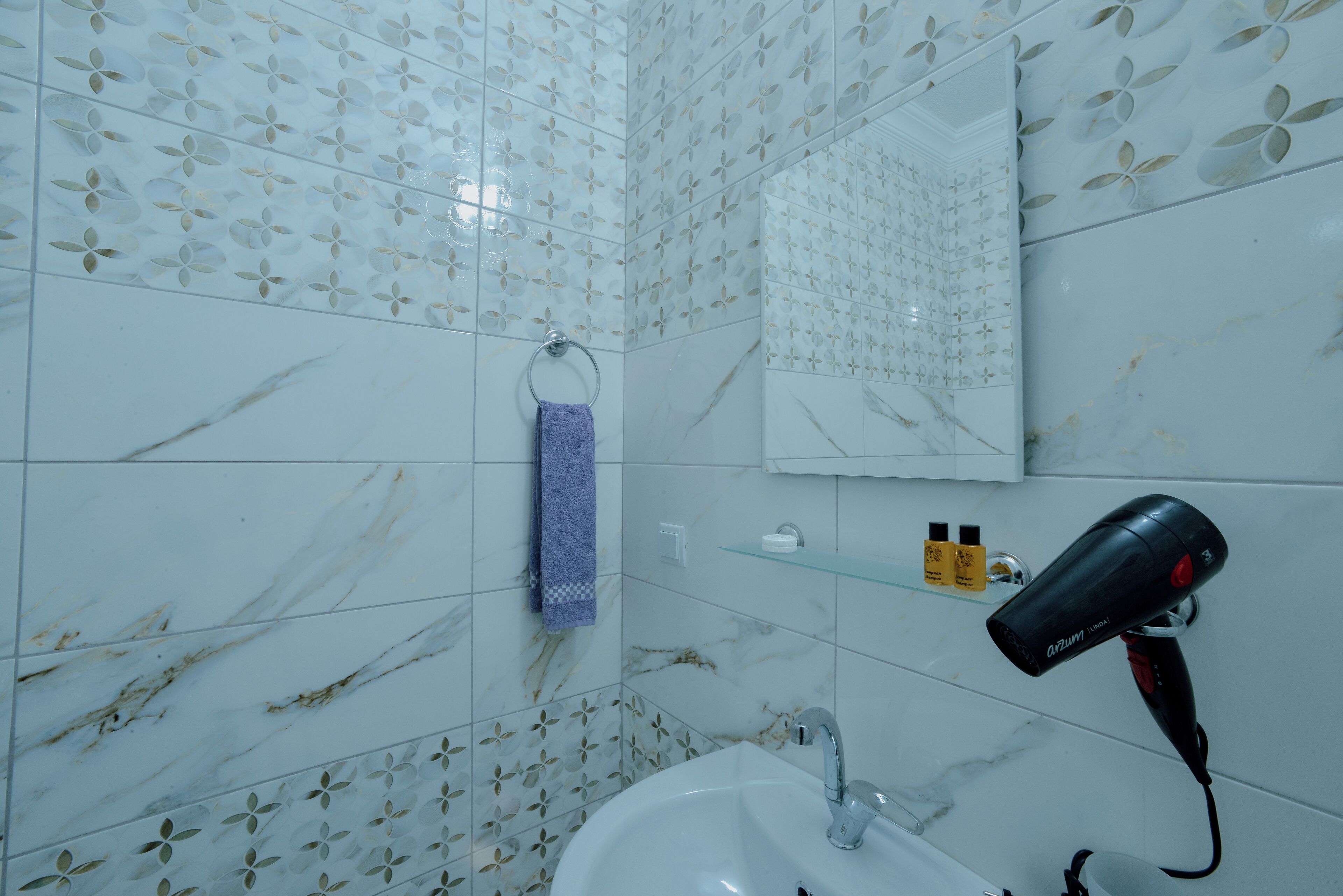 A bathroom with white marble tiles, a sink, a mirror, a towel ring, and a hairdryer on a shelf.