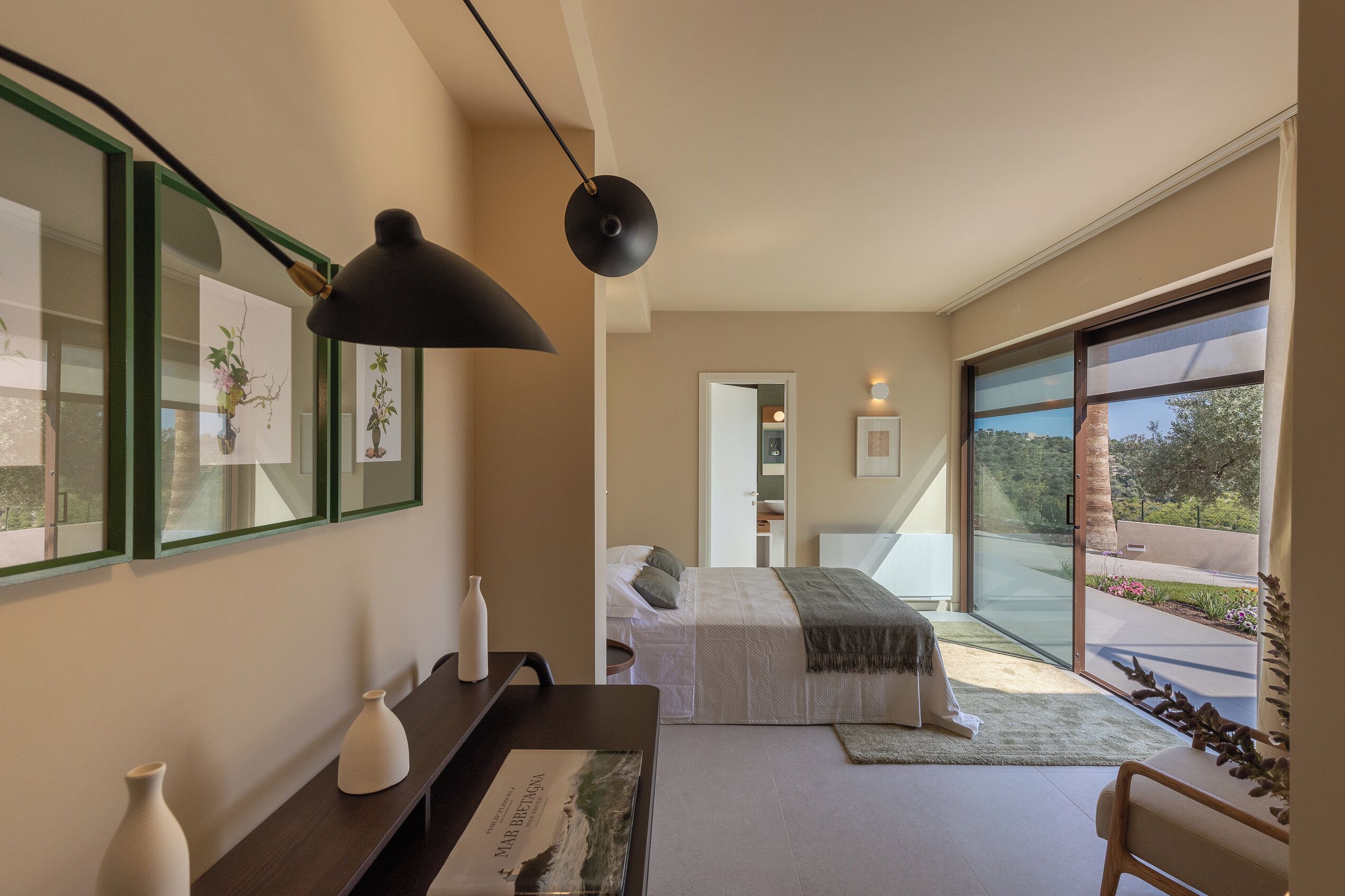 Une chambre à coucher moderne avec une grande porte-fenêtre coulissante donnant sur un balcon, un lit avec une tête de lit blanche et un bureau agrémenté d’un vase et d’un tableau encadré.