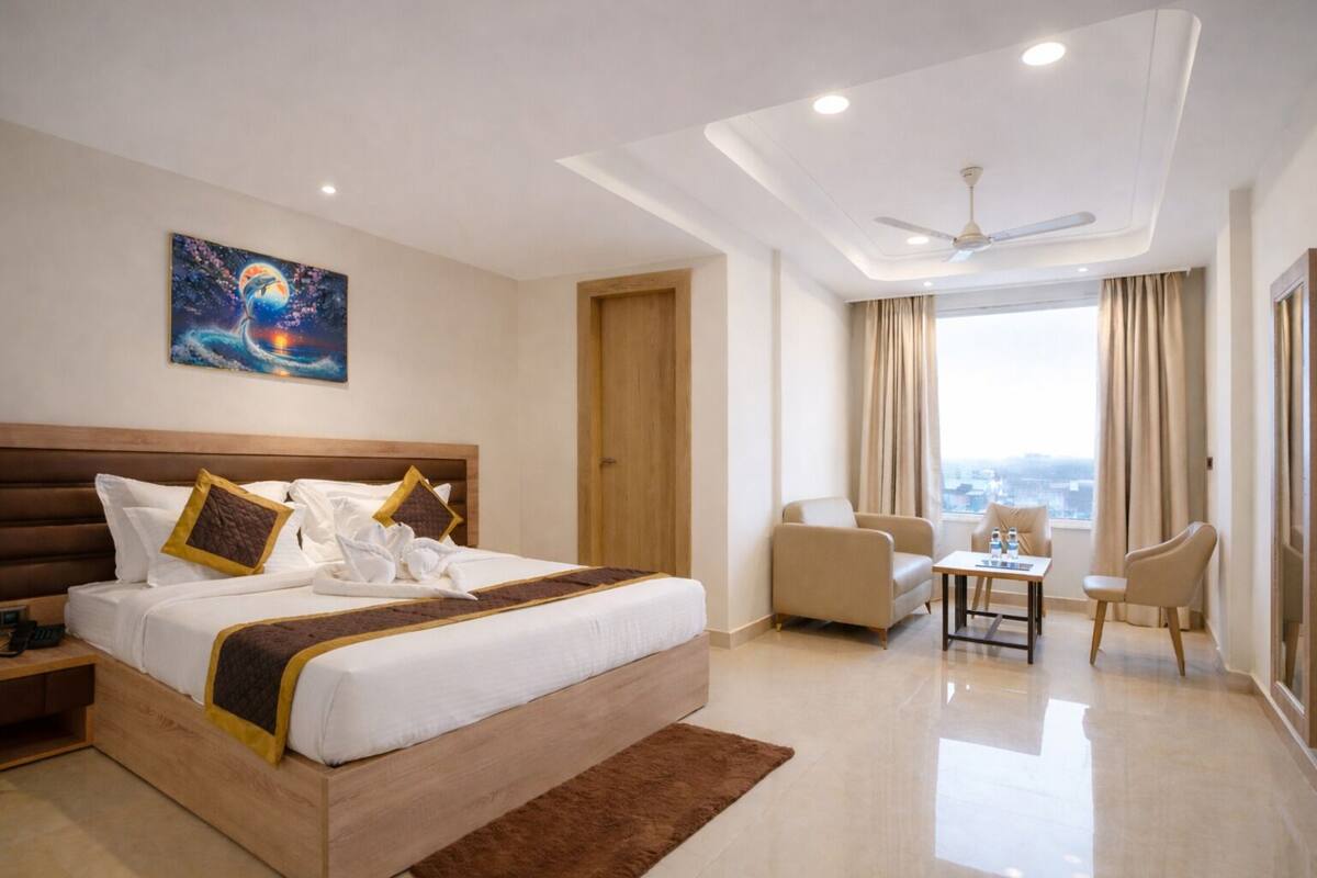 A hotel room with a large bed, a desk, two chairs, a ceiling fan, and a painting on the wall.