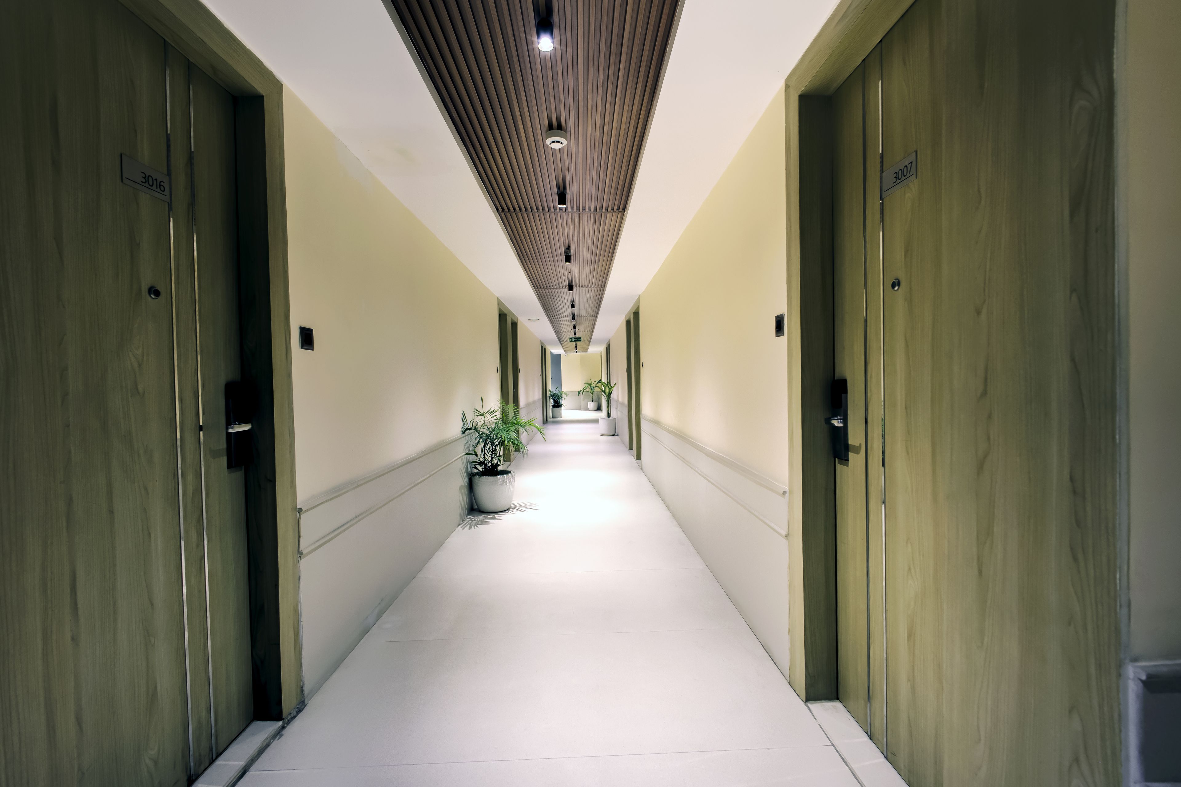 A hotel corridor with wooden doors, a white tiled floor, and a ceiling with recessed lighting.