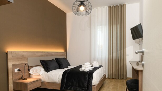 A modern hotel room with a wooden headboard, a bedside table with a lamp, a bed with white linens and black pillows, a black throw blanket, a desk with a monitor, and a large window with curtains. - L`Airone - Appartamento 2 (Campanarello)