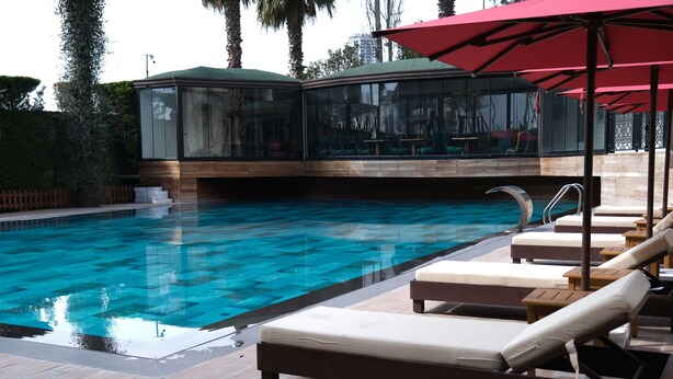 Una piscina con sillas de descanso y sombrillas, un área con paredes de vidrio y asientos, y una terraza de madera.