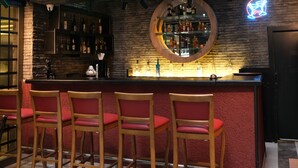 Zona de bar con letreros de neón, un espejo redondo y taburetes rojos.