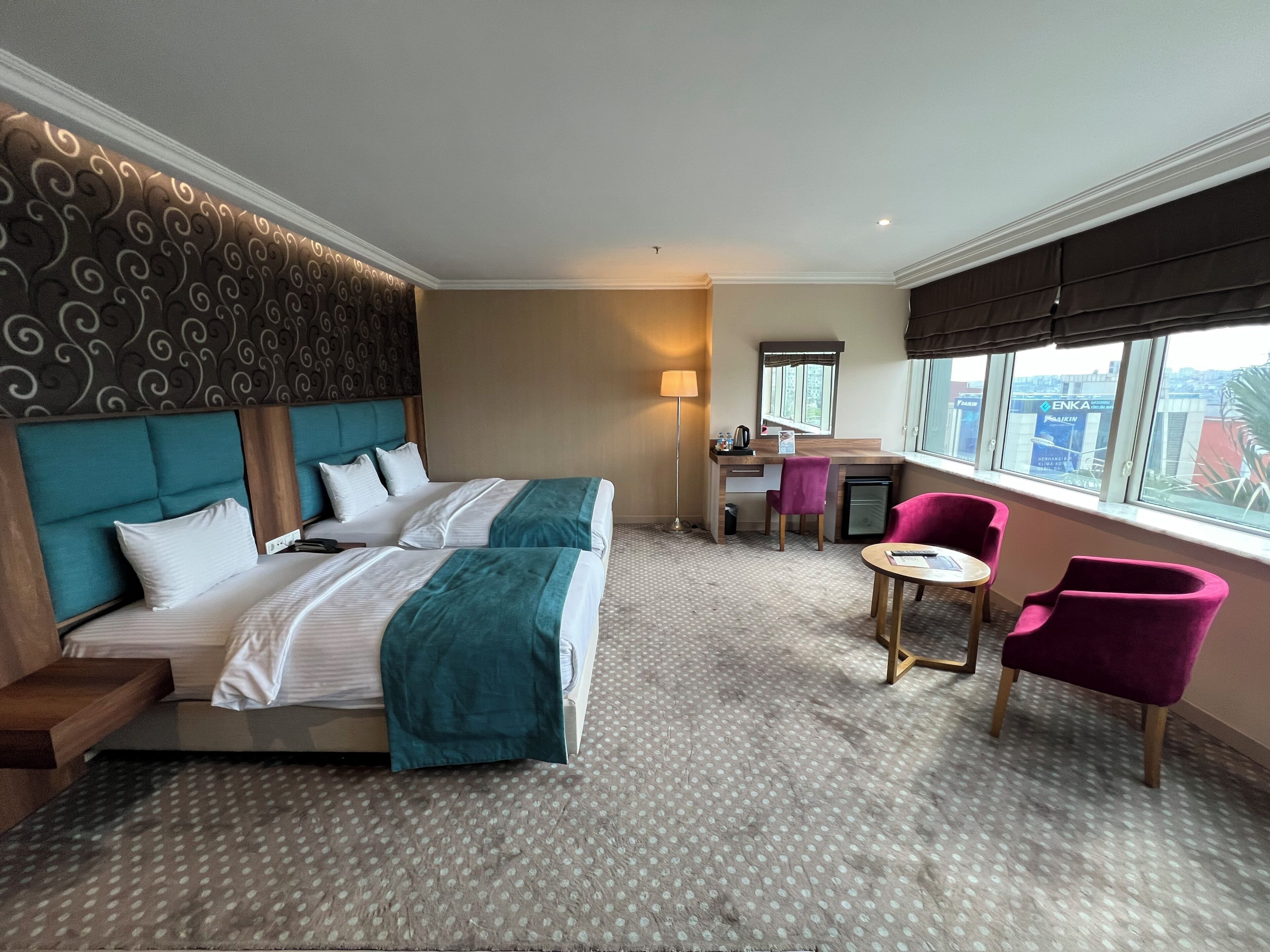 A hotel room with two beds, a desk, a chair, a small table, and a window with a city view.
