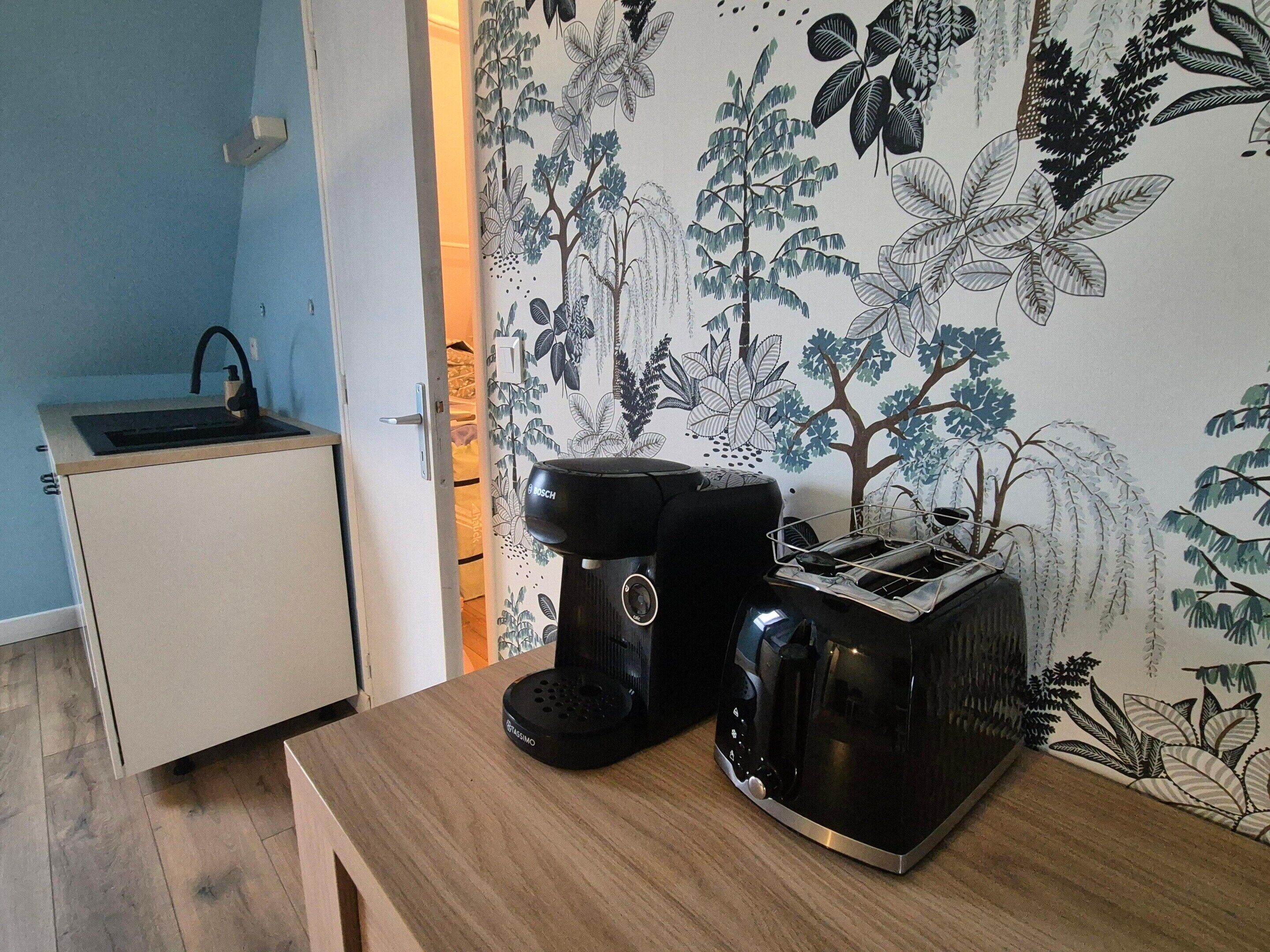 A kitchenette with a Bosch coffee maker and toaster on a wooden table, a sink, and a wall with a floral wallpaper pattern.