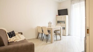 Ein Wohnzimmer mit einer Couch, einem Esstisch mit Stühlen, einem Fernseher auf einem Schrank und einem Fenster mit Vorhängen.