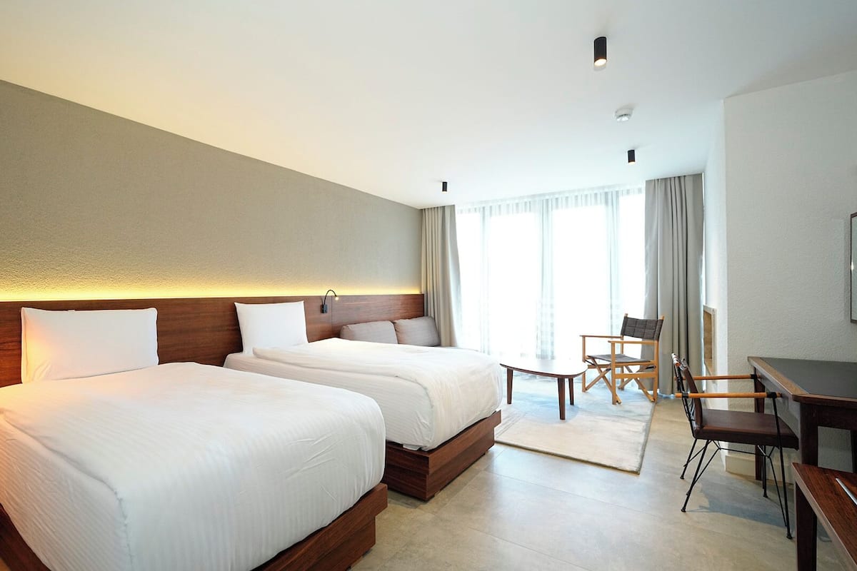 A hotel room with two beds, a desk, a chair, and a large window with curtains.