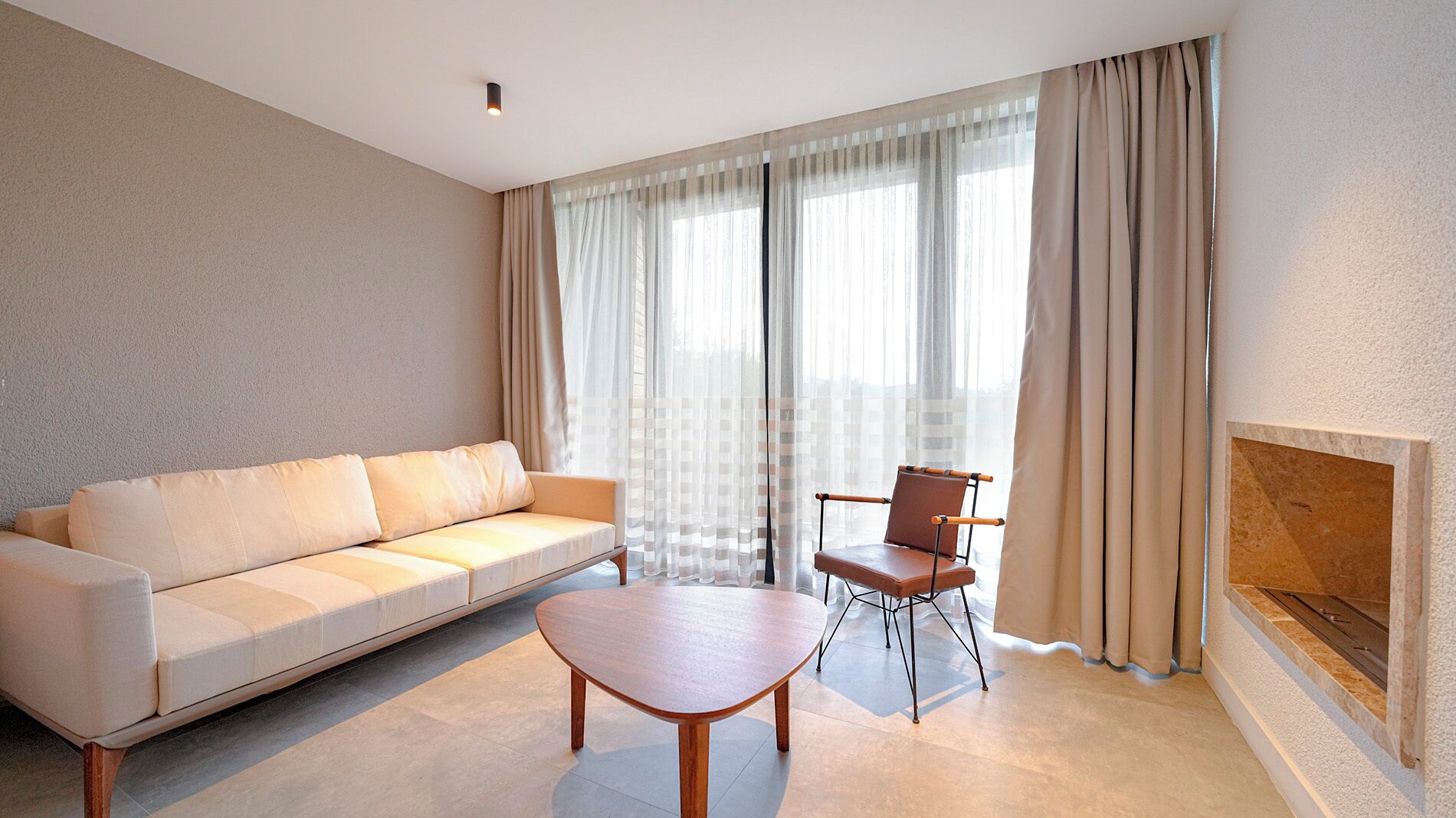 A modern living room with a sofa, a wooden coffee table, and a chair. There is a fireplace on the right and a large window with sheer curtains.